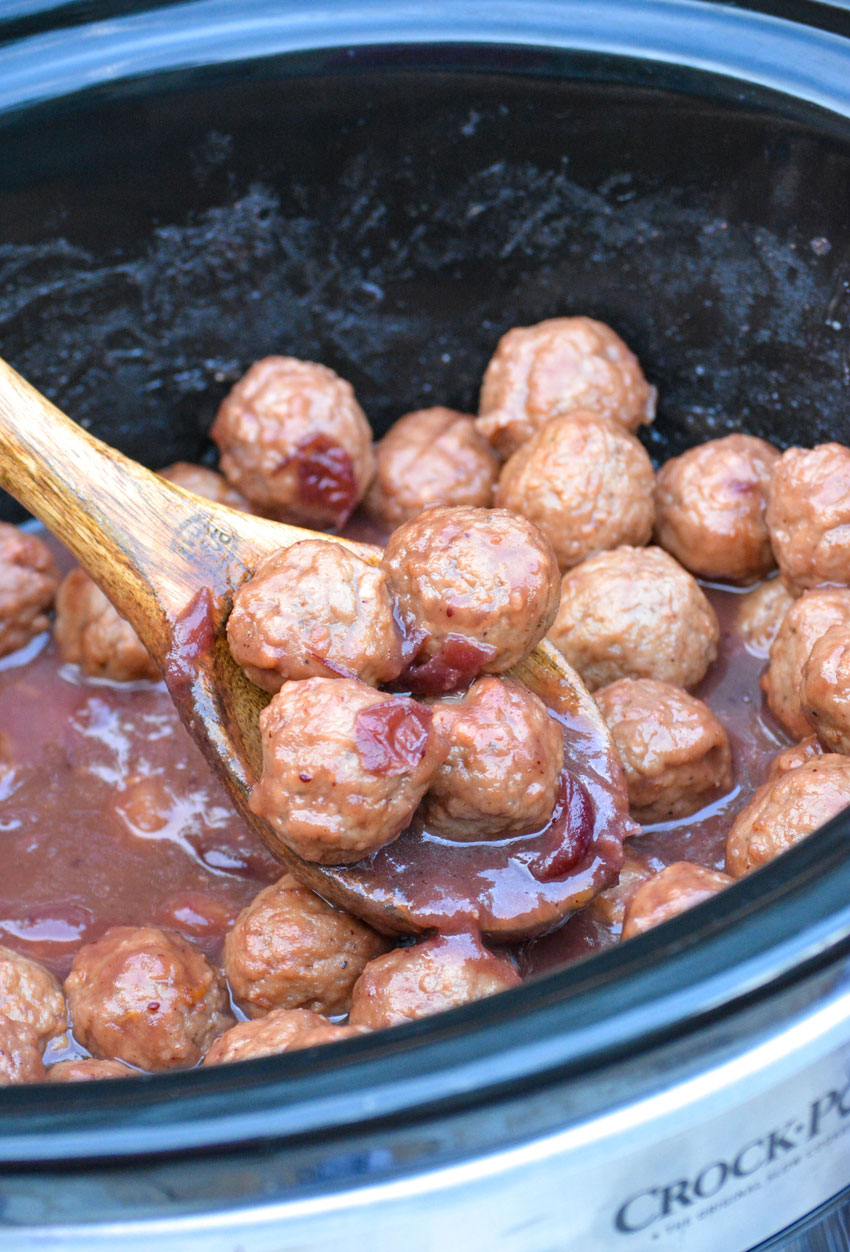 a wooden spoon shown holding four saucy cranberry meatballs above a black crockpot bowl