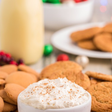 eggnog dip in a white bowl sprinkled with ground cinnamon and surrounded by vanilla wafers and gingersnap cookies