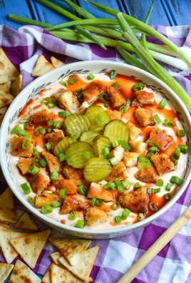 a shallow white serving bowl filled with nashville hot chicken dip surrounded by green onions and triangle shaped crackers