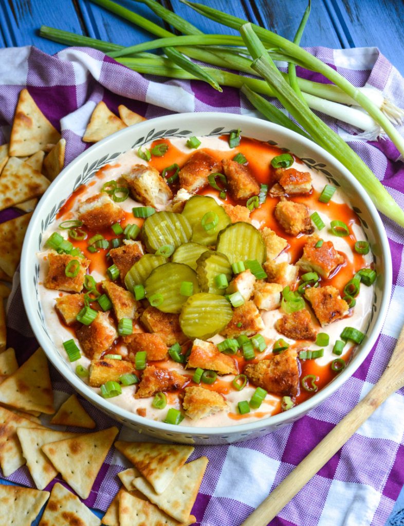 a shallow white serving bowl filled with nashville hot chicken dip surrounded by green onions and triangle shaped crackers