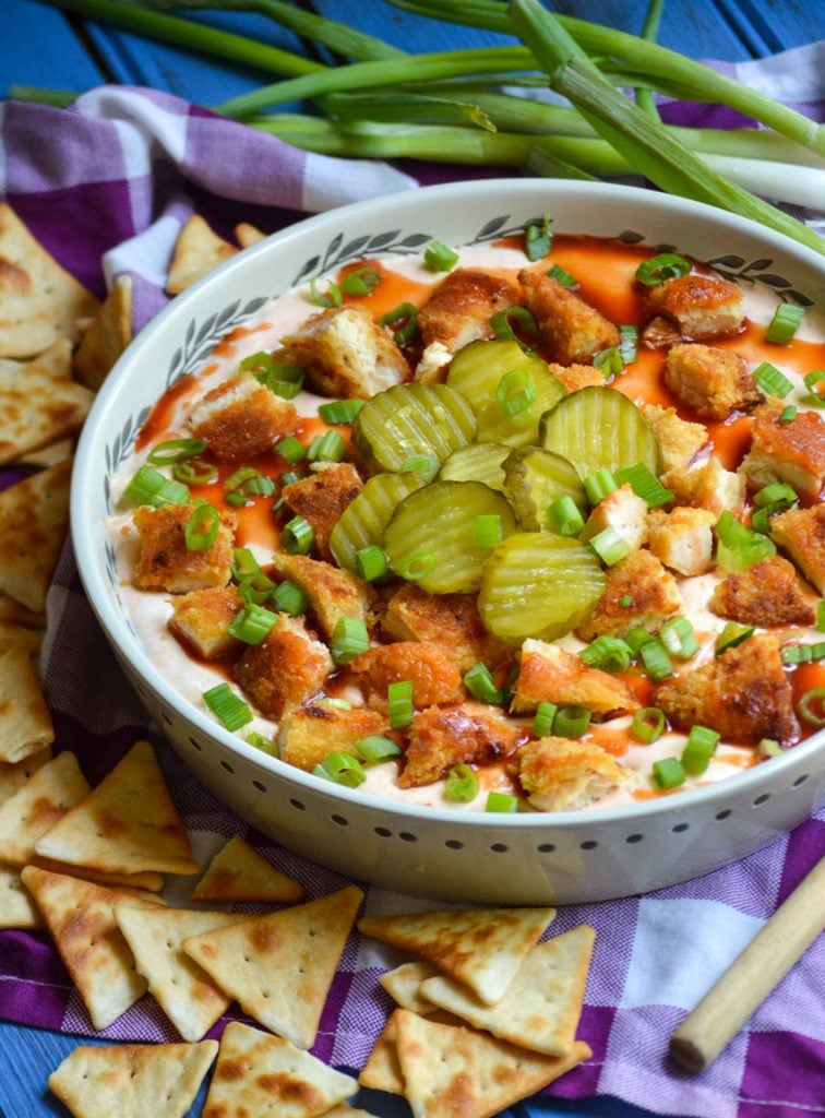 a shallow white serving bowl filled with nashville hot chicken dip surrounded by green onions and triangle shaped crackers