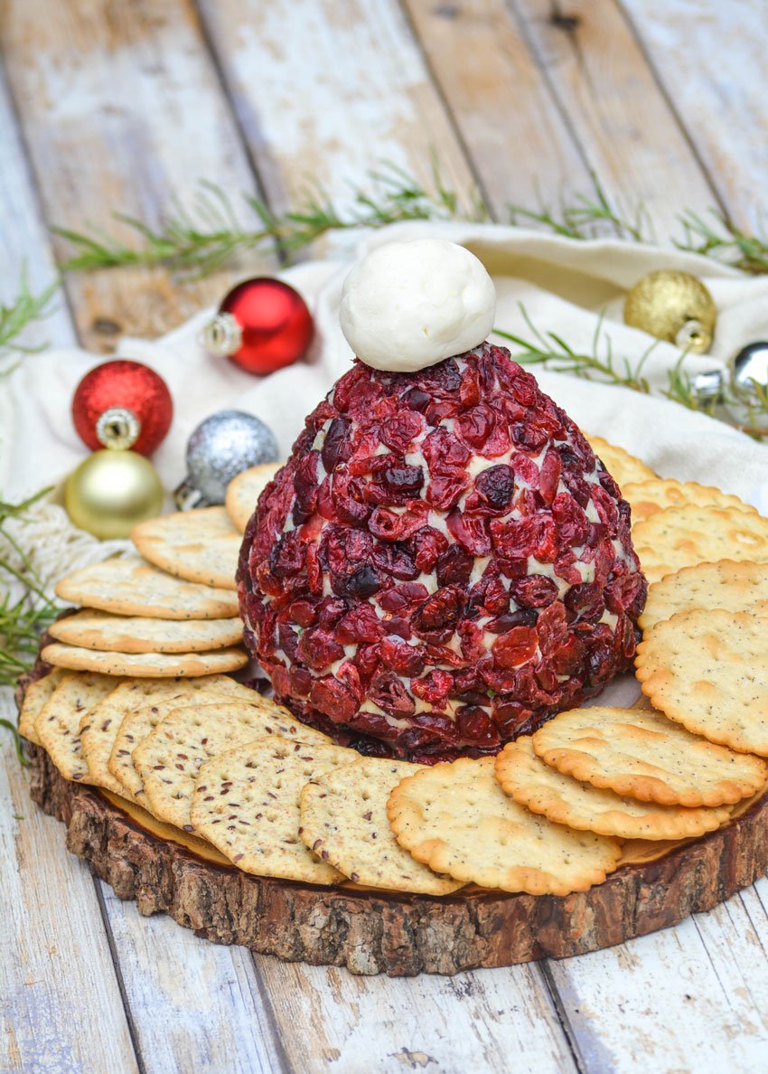 cranberry cheddar santa hat cheeseball surrounded by crackers with small ornaments & sprigs of rosemary in the background