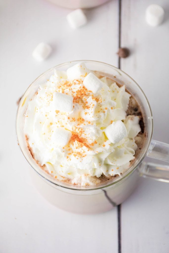 a mug of eggnog hot cocoa topped with whipped cream, golden sprinkles, and mini marshmallows