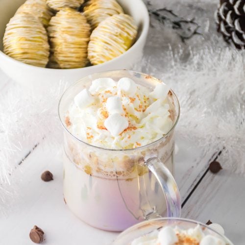 eggnog hot cocoa bombs in a white bowl behind two glass mugs filled with whip cream topped cocoa