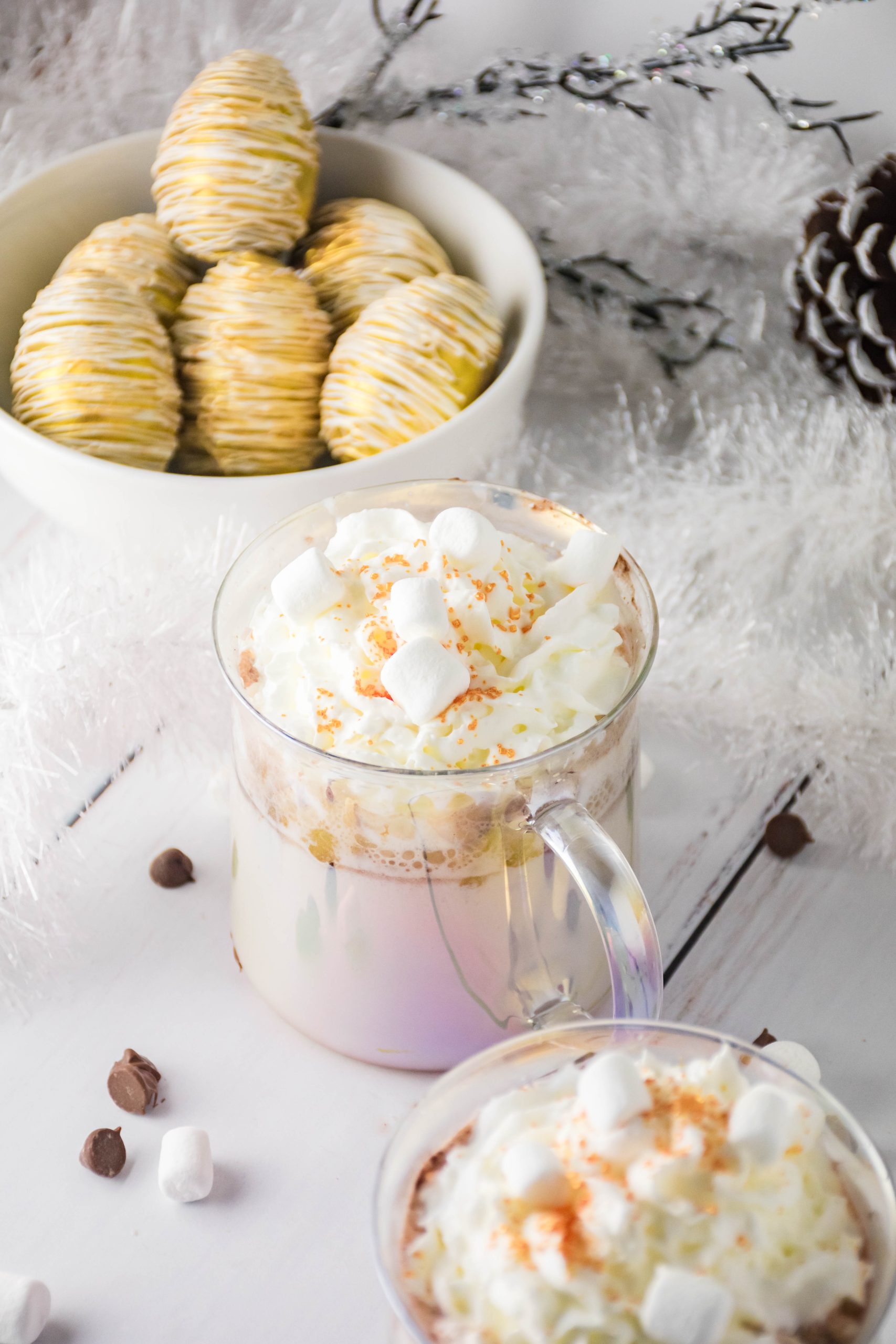 eggnog hot cocoa bombs in a white bowl behind two glass mugs filled with whip cream topped cocoa