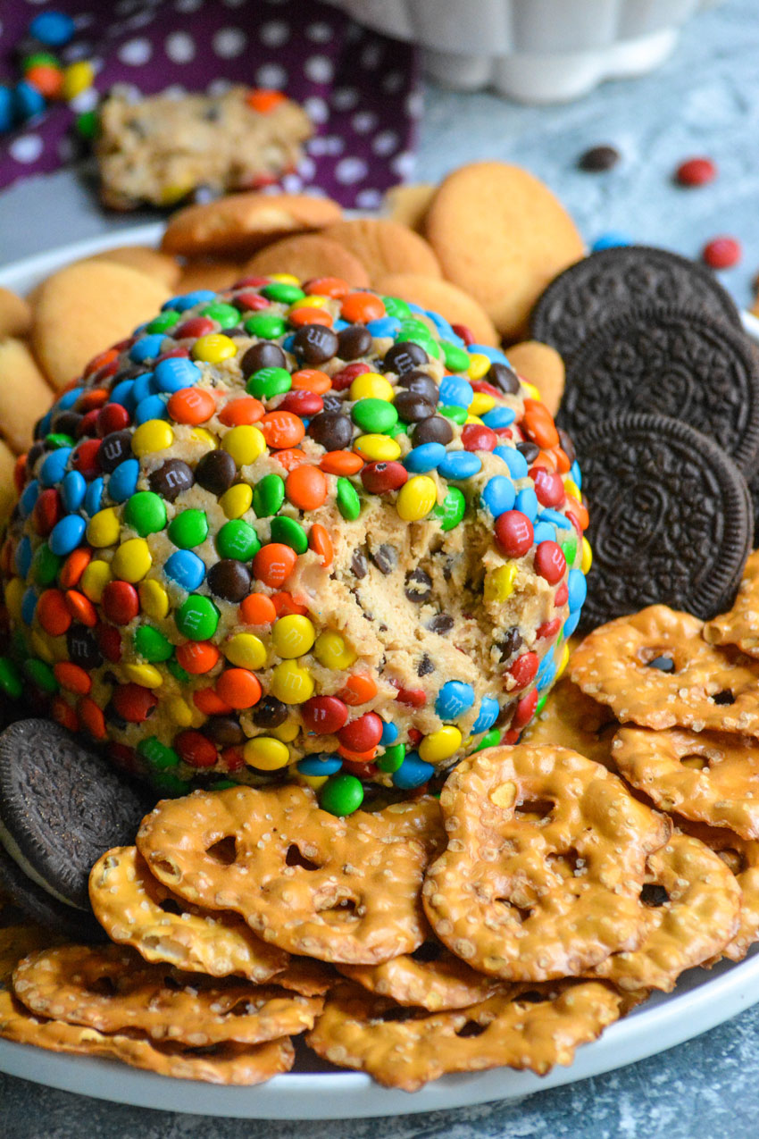 a monster cookie cheeseball on a white plate surrounded by pretzels and cookies