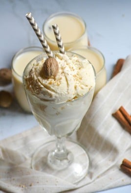 a glass filled with scoops of no churn eggnog ice cream topped with golden sugar sprinkles and freshly grated nutmeg