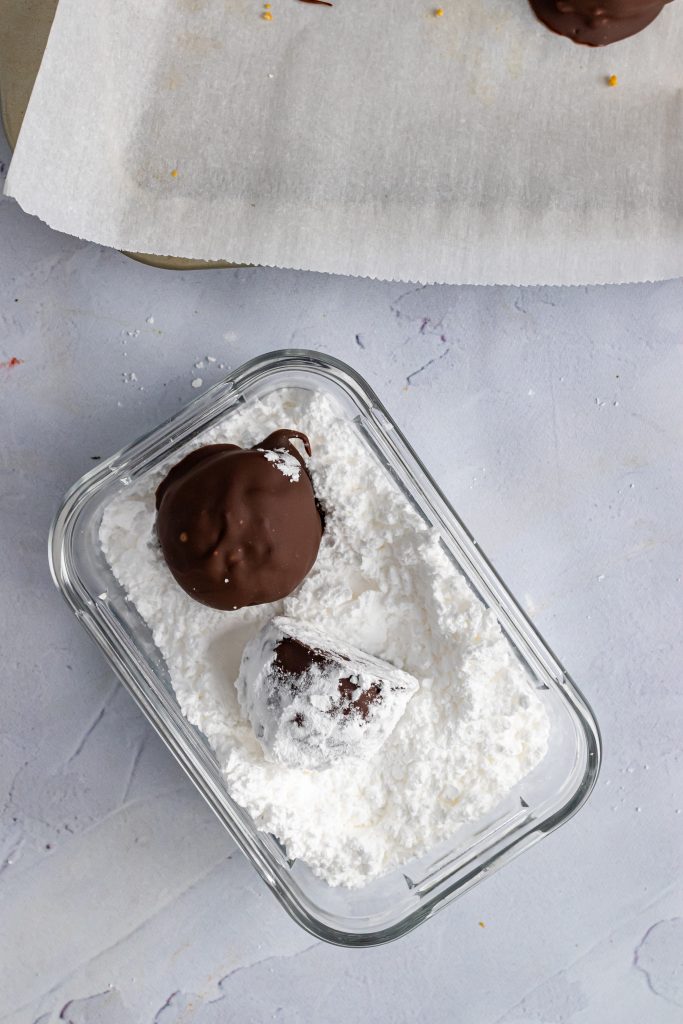 a puppy chow truffle ball being rolled in a bowl of fluffy white powdered sugar
