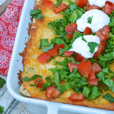 Mexican lasagna in a white casserole dish topped with lettuce, tomatoes, and dollops of sour cream
