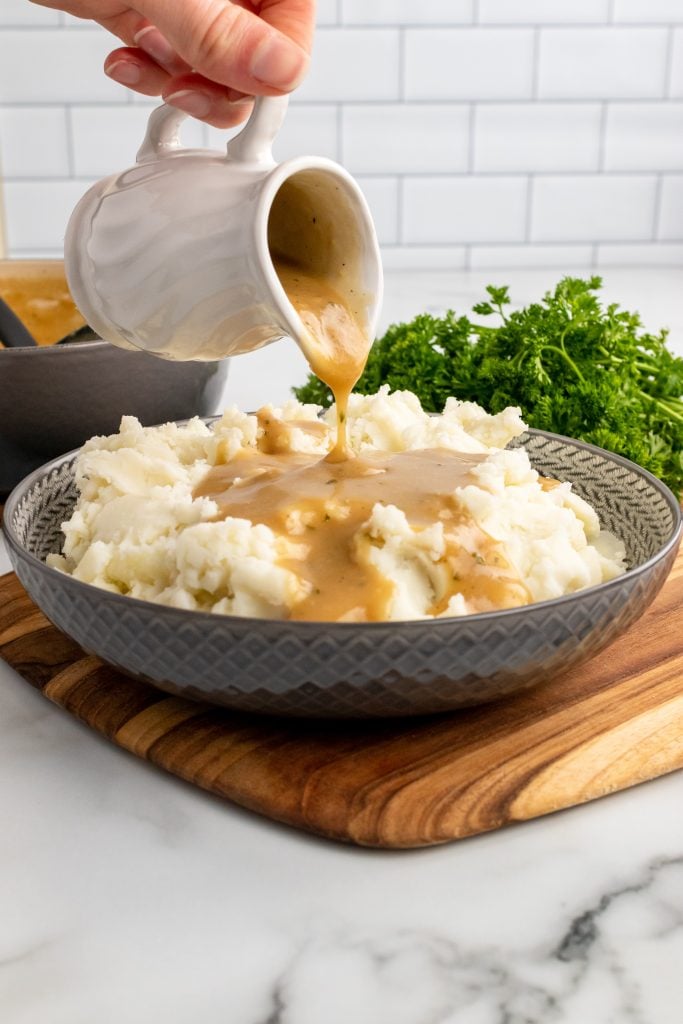 a small white pitcher filled with easy homemade brown gravy being poured over a gray bowl filled with fluffy mashed potatoes