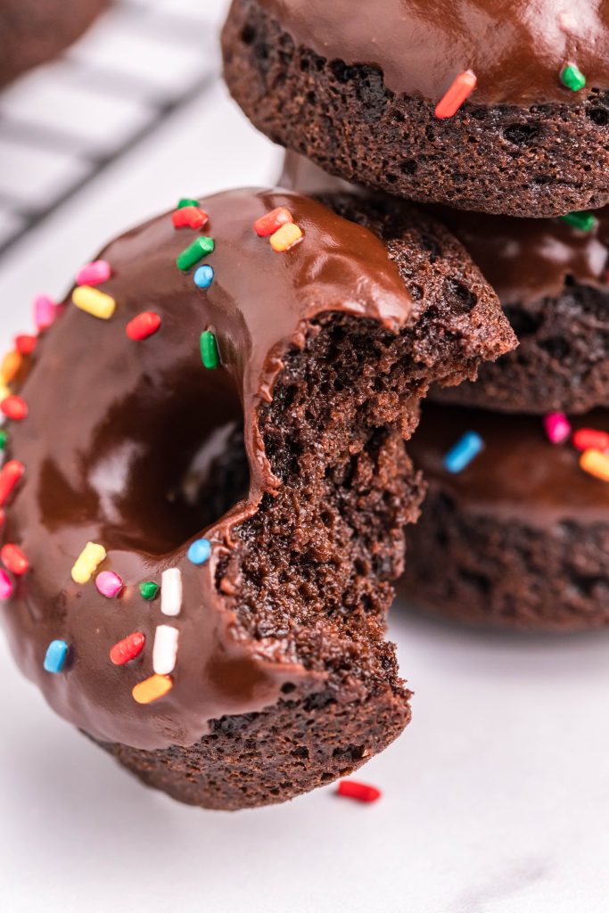 a double chocolate donut with a bite taken out of it resting against a stack of chocolate donuts