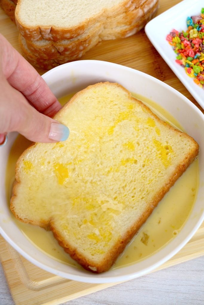 a piece of bread being dipped in egg wash in a white bowl