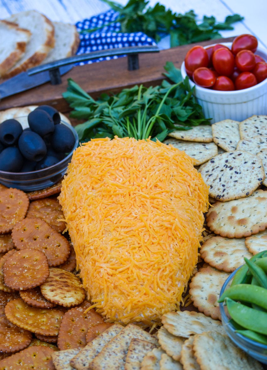 carrot shaped cheddar cheese ball on a wooden serving tray surrounded by crackers and fresh vegetables