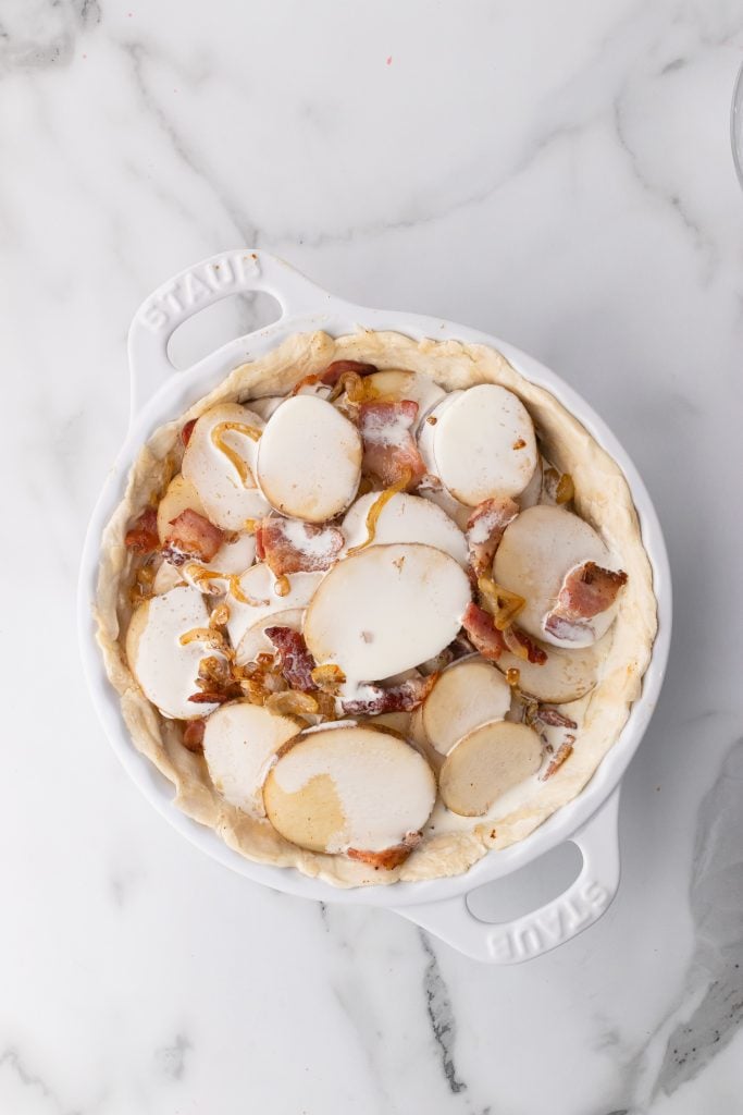sliced potatoes, caramelized onion and crisp bacon layered in a pie crust in a white dish and topped with cream
