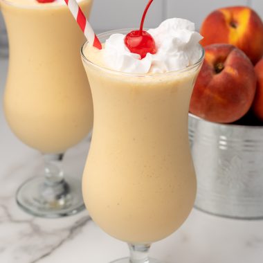 copycat chick fil a peach milkshake in two large glasses topped with whipped cream and a maraschino cherry on top