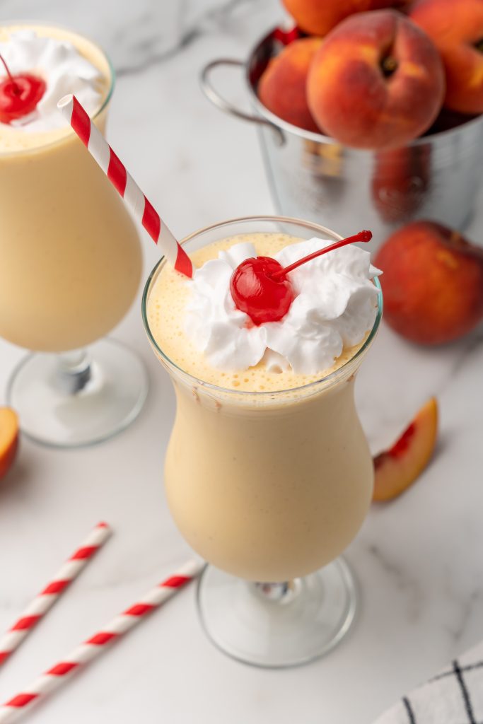 copycat chick fil a peach milkshake in two large glasses topped with whipped cream and a maraschino cherry on top
