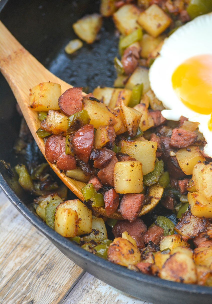 a wooden spoon in a skillet of crispy potato hash with peppers, onions, and chopped hot dogs