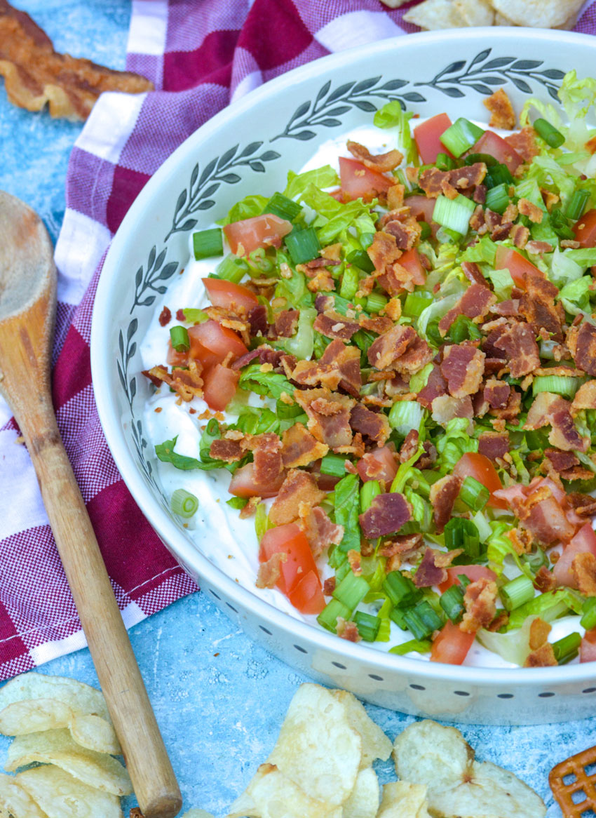 creamy blt dip in a shallow serving bowl topped with shredded lettuce, crumbled bacon, diced tomatoes, and thinly sliced green onions
