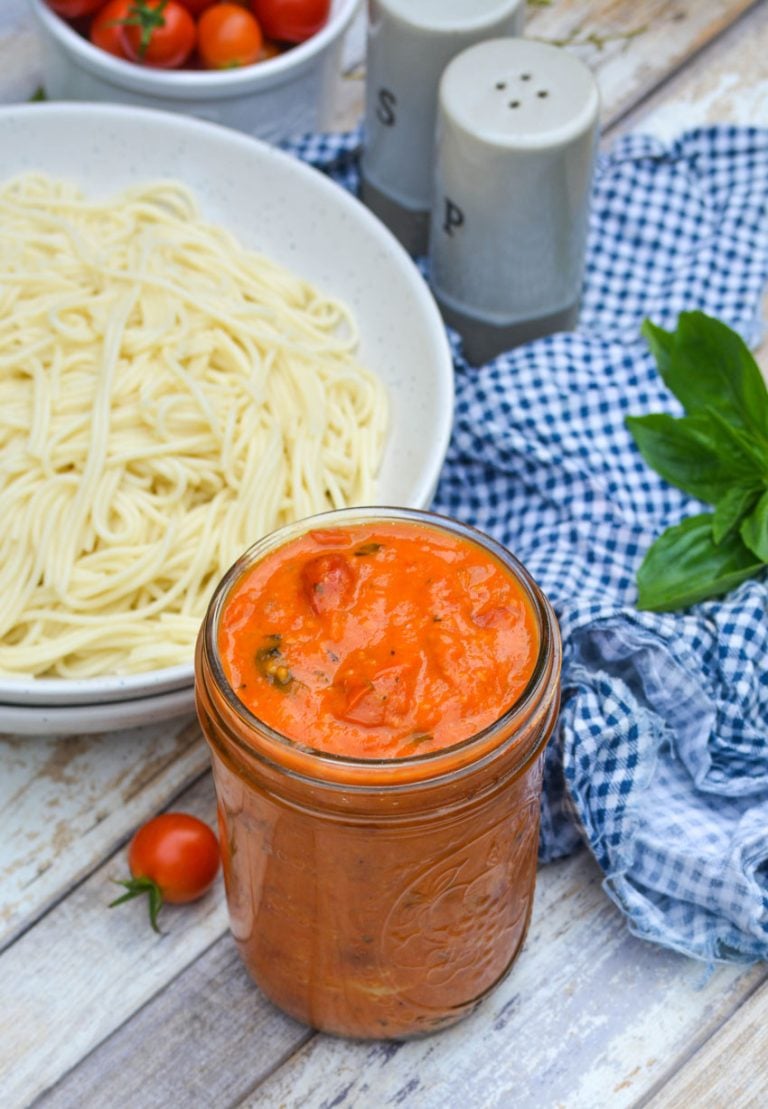 sweet cherry tomato sauce in a glass jar in front of a white bowl filled with cooked spaghetti