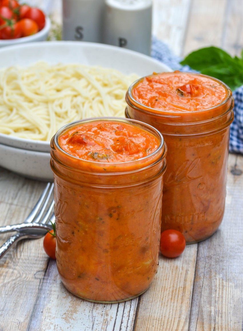 sweet cherry tomato sauce in a glass jar in front of a white bowl filled with cooked spaghetti