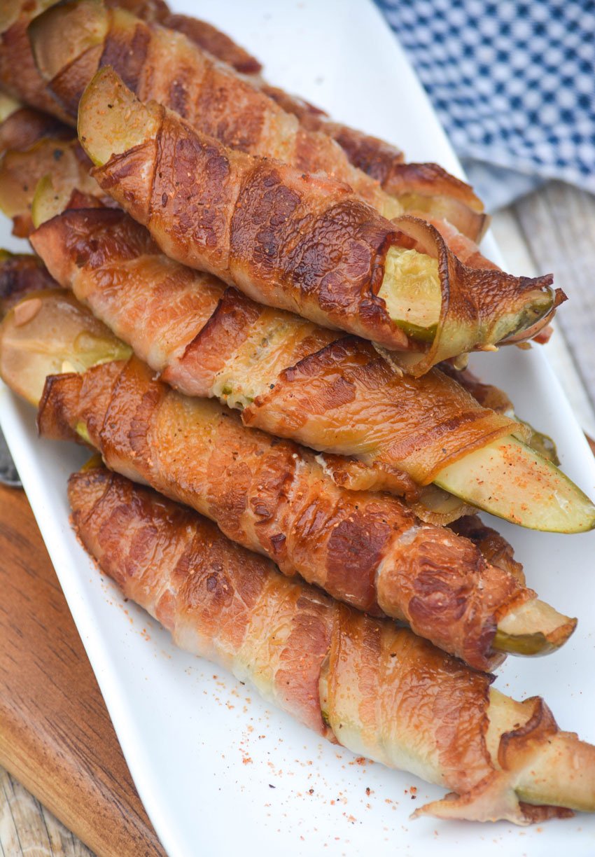 a stacked pile of bacon wrapped smoked pickles on a white serving platter