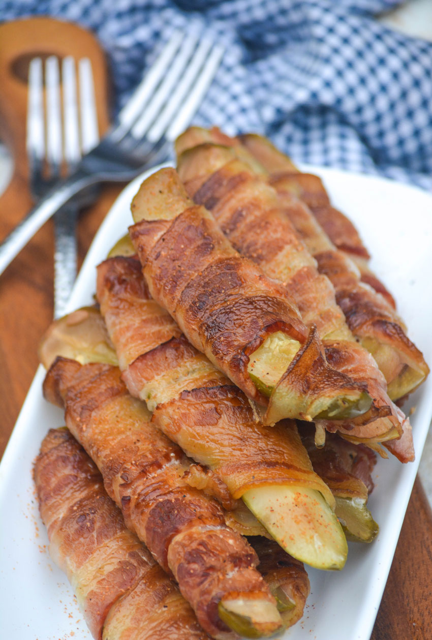 a stacked pile of bacon wrapped smoked pickles on a white serving platter