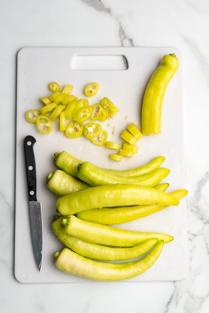 whole pickled banana peppers and some slices on a white cutting board with a small black handled paring knife