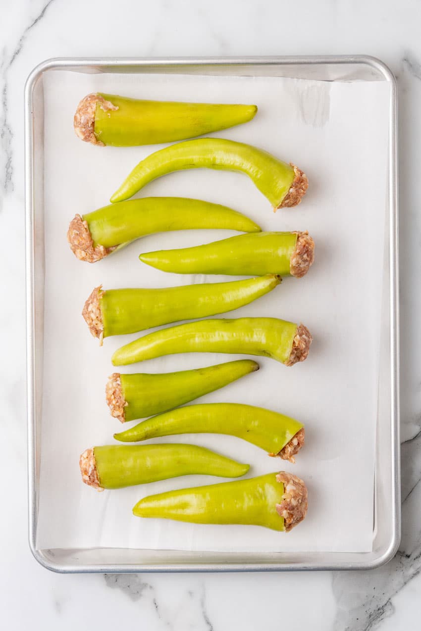 unbaked sausage stuffed banana peppers arranged in a row on a parchment paper lined baking sheet