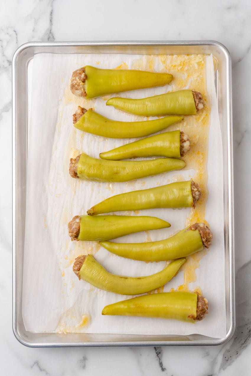 baked sausage stuffed banana peppers arranged in a row on a parchment paper lined baking sheet