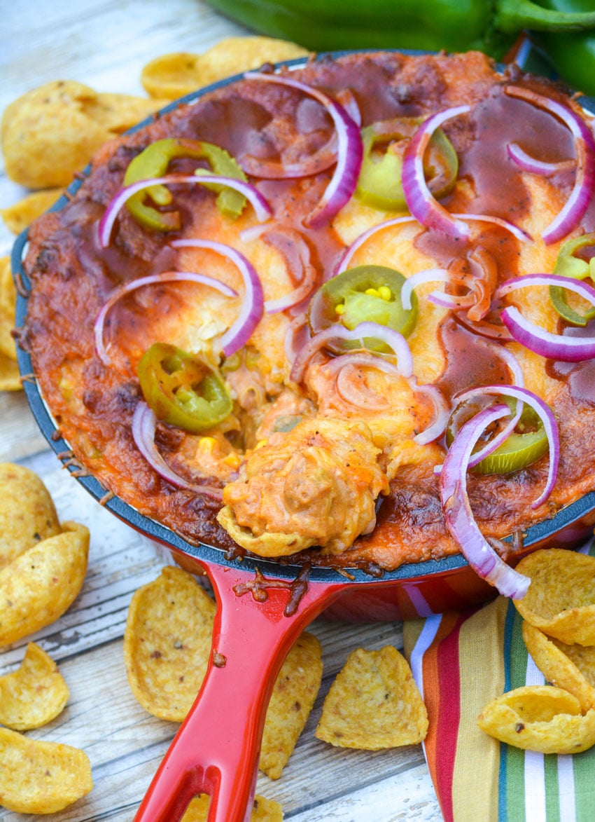 a corn chip topped with cheesy pulled pork barbecue dip resting on the edge of a small red skillet