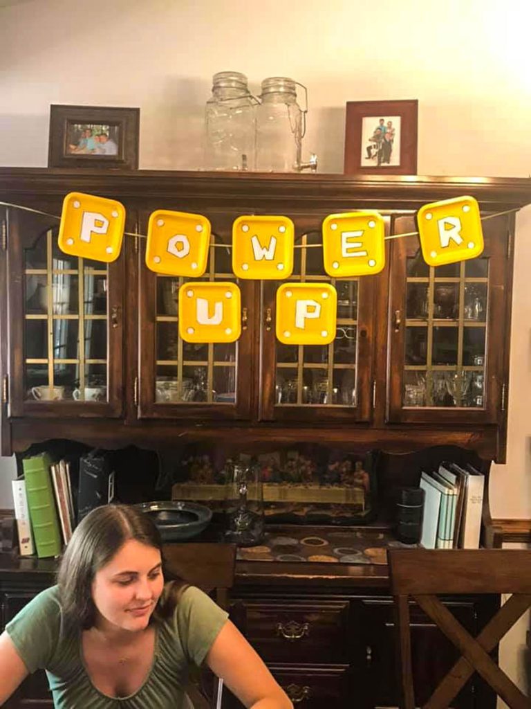 homemade mario power up banner made with square yellow paper plates hung from a brown china hutch