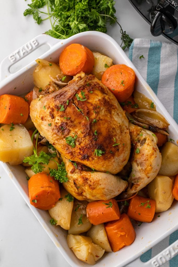 slow cooker whole chicken in a white dish resting on a bed of tender potatoes and carrots