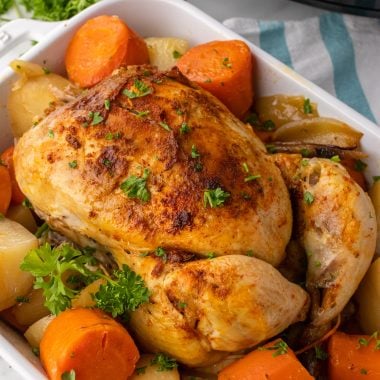 slow cooker whole chicken in a white dish resting on a bed of tender potatoes and carrots