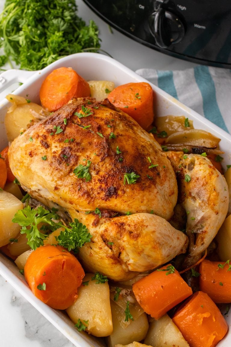 slow cooker whole chicken in a white dish resting on a bed of tender potatoes and carrots