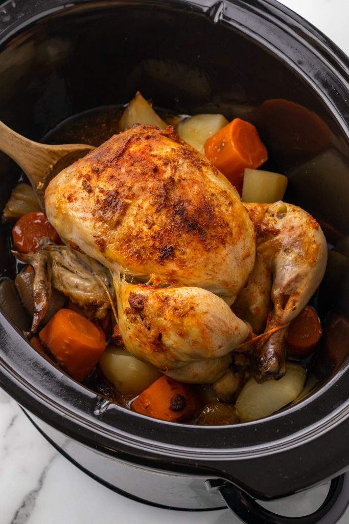 slow cooker whole chicken in a black crockpot on top of sliced carrots and potatoes