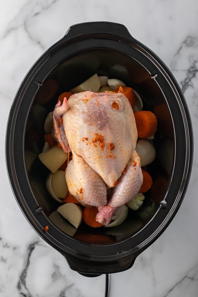 a raw whole chicken resting on a bed of cut vegetables in a black crockpot
