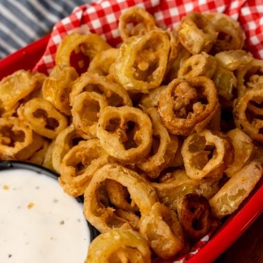 deep fried banana peppers in a red basket lined with checkered paper with a bowl of ranch for dipping