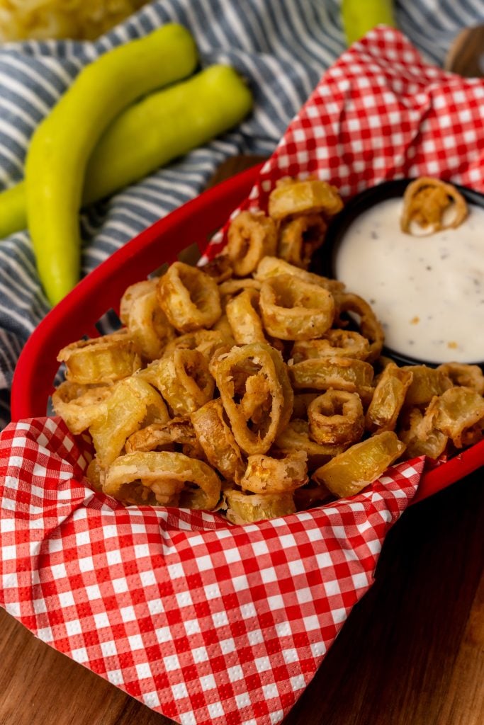 deep fried banana peppers in a red basket lined with checkered paper with a bowl of ranch for dipping
