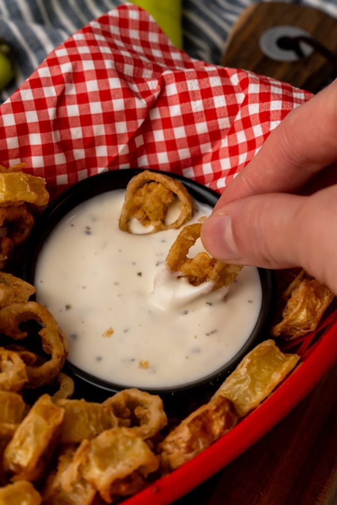 a hand dipping fried banana pepper rings into a black bowl filled with creamy ranch dressing