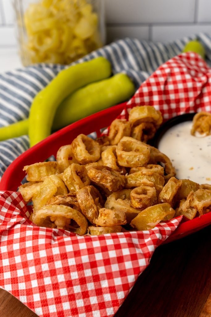 deep fried banana peppers in a red basket lined with checkered paper with a bowl of ranch for dipping