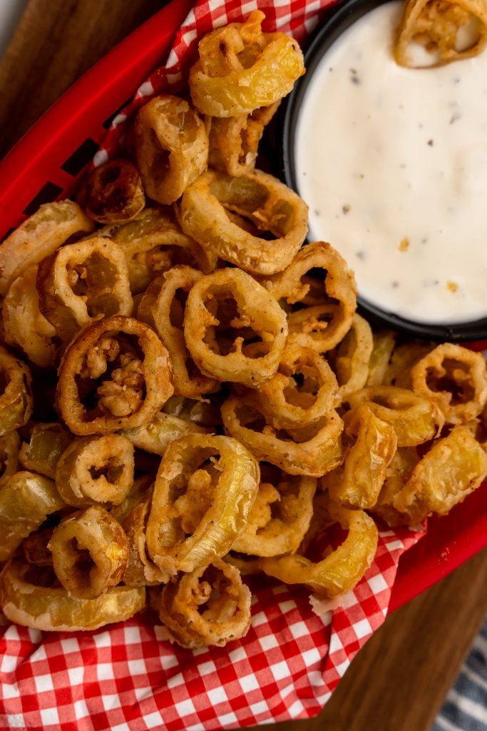 deep fried banana peppers in a red basket lined with checkered paper with a bowl of ranch for dipping