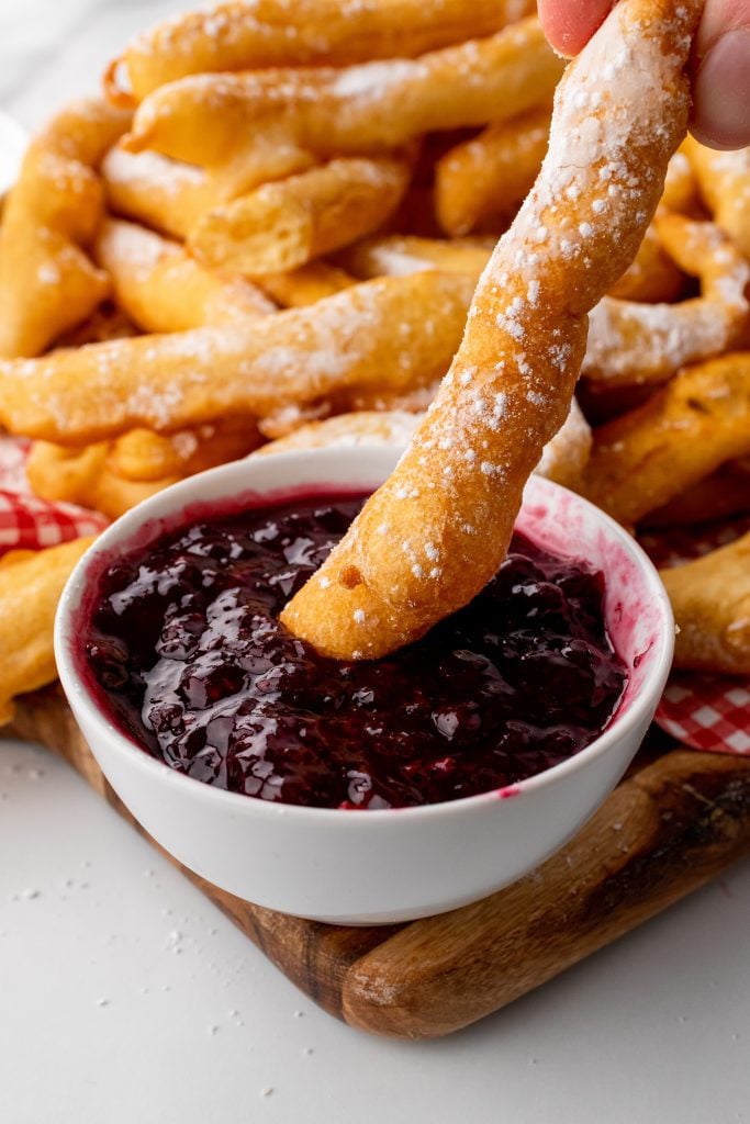 a funnel cake fry being dunked in a small white bowl filled with raspberry jam