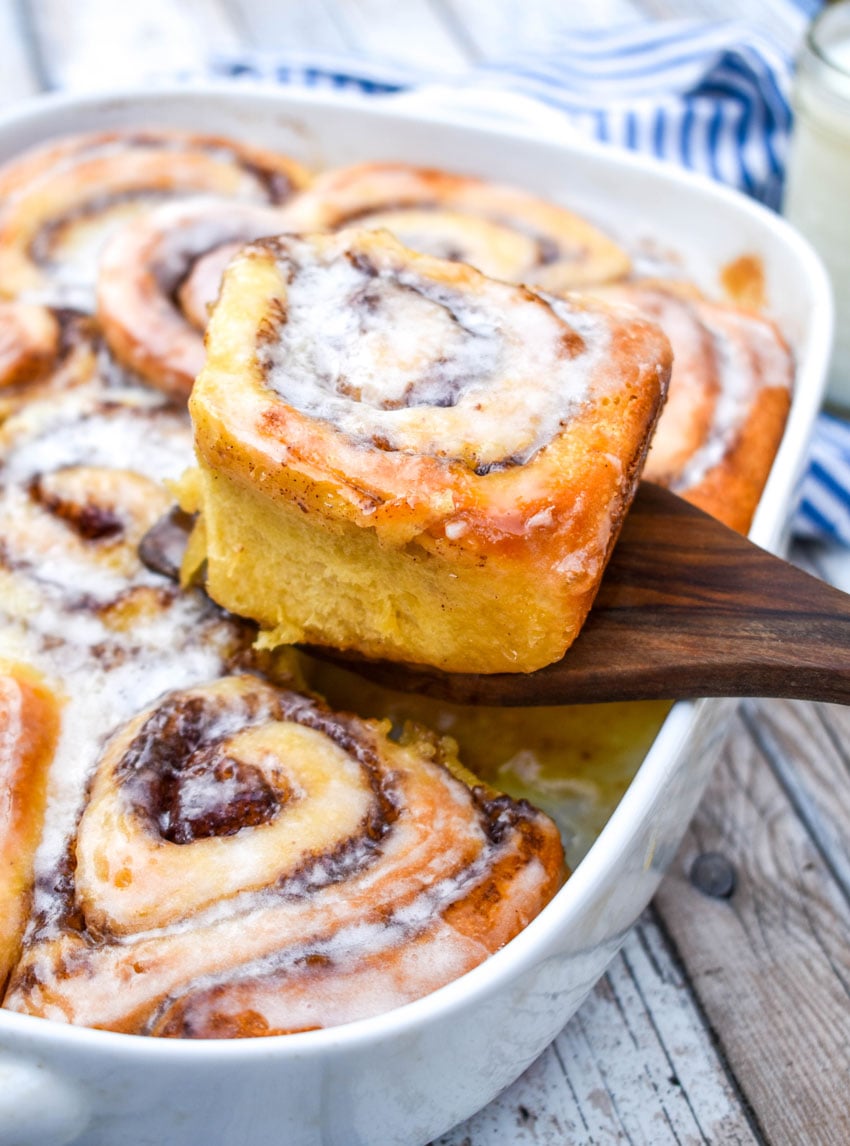 a wooden spatula lifting up one of tiktok cinnamon rolls with heavy cream out of a white baking dish