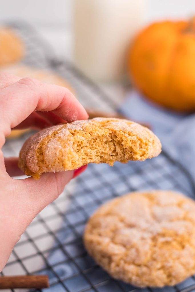 a hand holding up a pumpkin sugar cookie with a bite taken out of the center