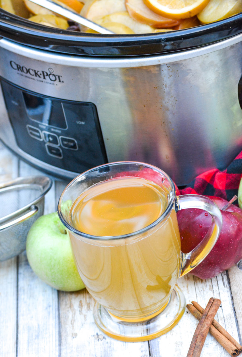 a clear glass filled with homemade apple cider in front of a silver slow cooker