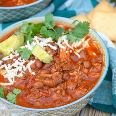 Slow cooker pulled pork chili in a blue bowl topped with cheese, cilantro leaves, and chunks of avocado