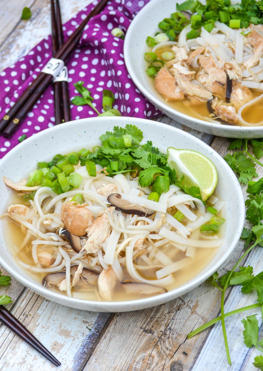 two bowls of easy crockpot chicken pho topped with cilantro leaves and lime wedges with a pair of chop sticks on the side