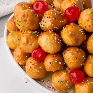 Nonna's Italian struffoli balls on a white platter sprinkled with sprinkles and maraschino cherries