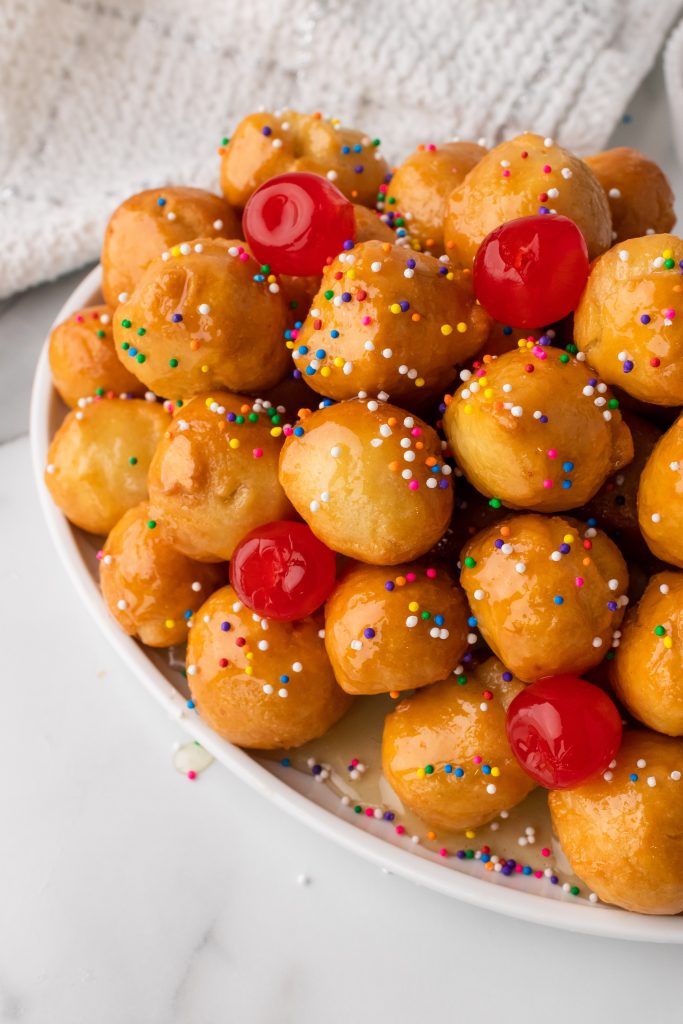 Nonna's Italian struffoli balls on a white platter sprinkled with sprinkles and maraschino cherries