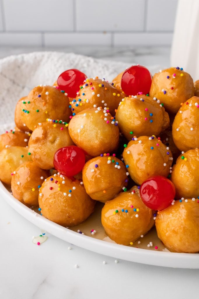 Nonna's Italian struffoli balls on a white platter sprinkled with sprinkles and maraschino cherries